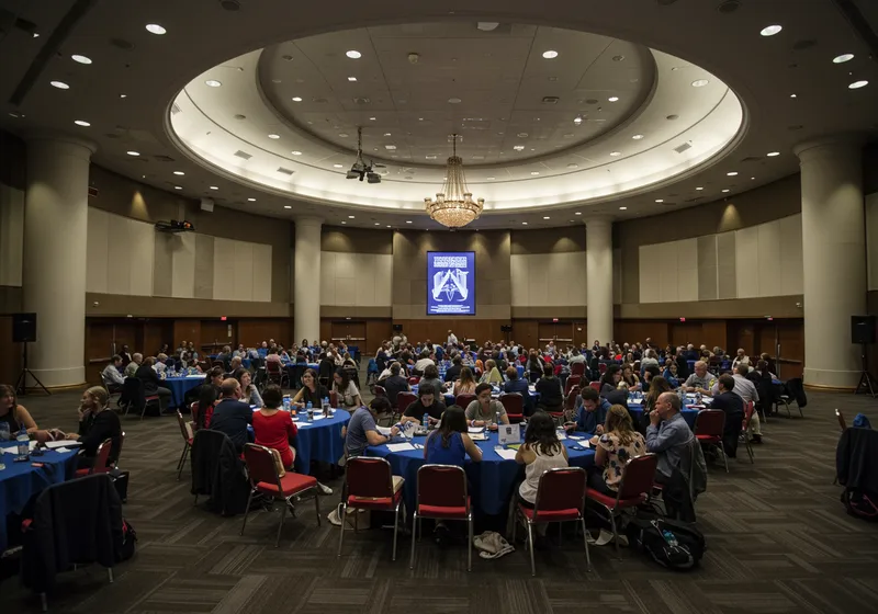 A busy symposium at the Galveston Island Convention Center, with professionals seated at round tables, engaging with laptops, documents, and group discussions.